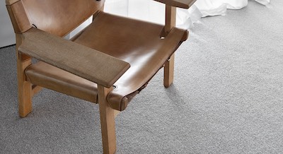 A rustic chair made of wood and brown leather, sitting near a window in a room laid with Crosbie 132 from the Boden carpet range.