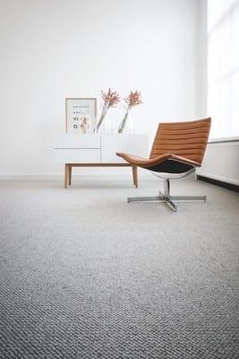 A creamy-coloured loop-pile carpet laid in a room with white walls and a brown Eames-inspired seat.