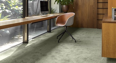 A mid-century modern style home office, brightly lit with natural light from a floor-to-ceiling window, contrasts wooden furniture with the green hue of Mezen, a plush nylon carpet from the Mink range.
